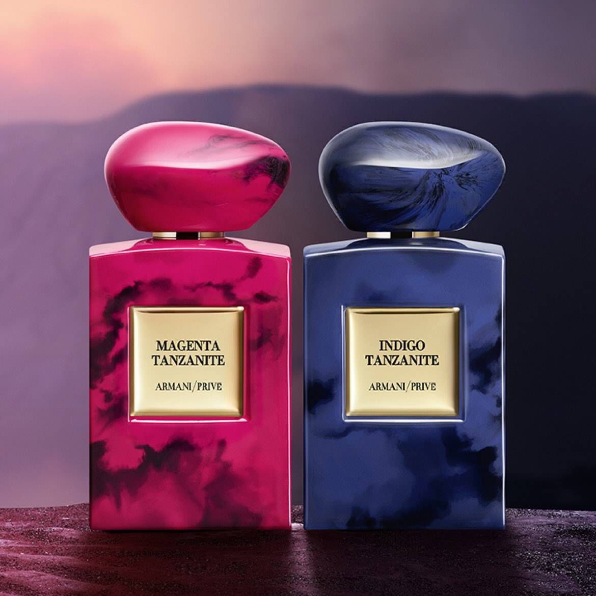 Two elegantly designed perfume bottles side by side: one is vibrant magenta and the other deep indigo, both featuring a smooth, rounded cap and gold-label branding. The backdrop suggests a dramatic landscape, enhancing the luxurious feel of the bottles.
