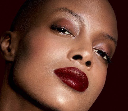 A close-up portrait of a woman with a shaved head, showcasing her striking features and bold, deep red lips. She wears a sparkling, embellished dress and looks confidently at the camera with a slight tilt of her head against a dark background.