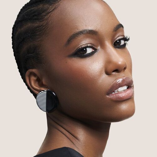 A close-up portrait of a woman with braided hair and bold makeup, featuring large striped earrings against a neutral background.