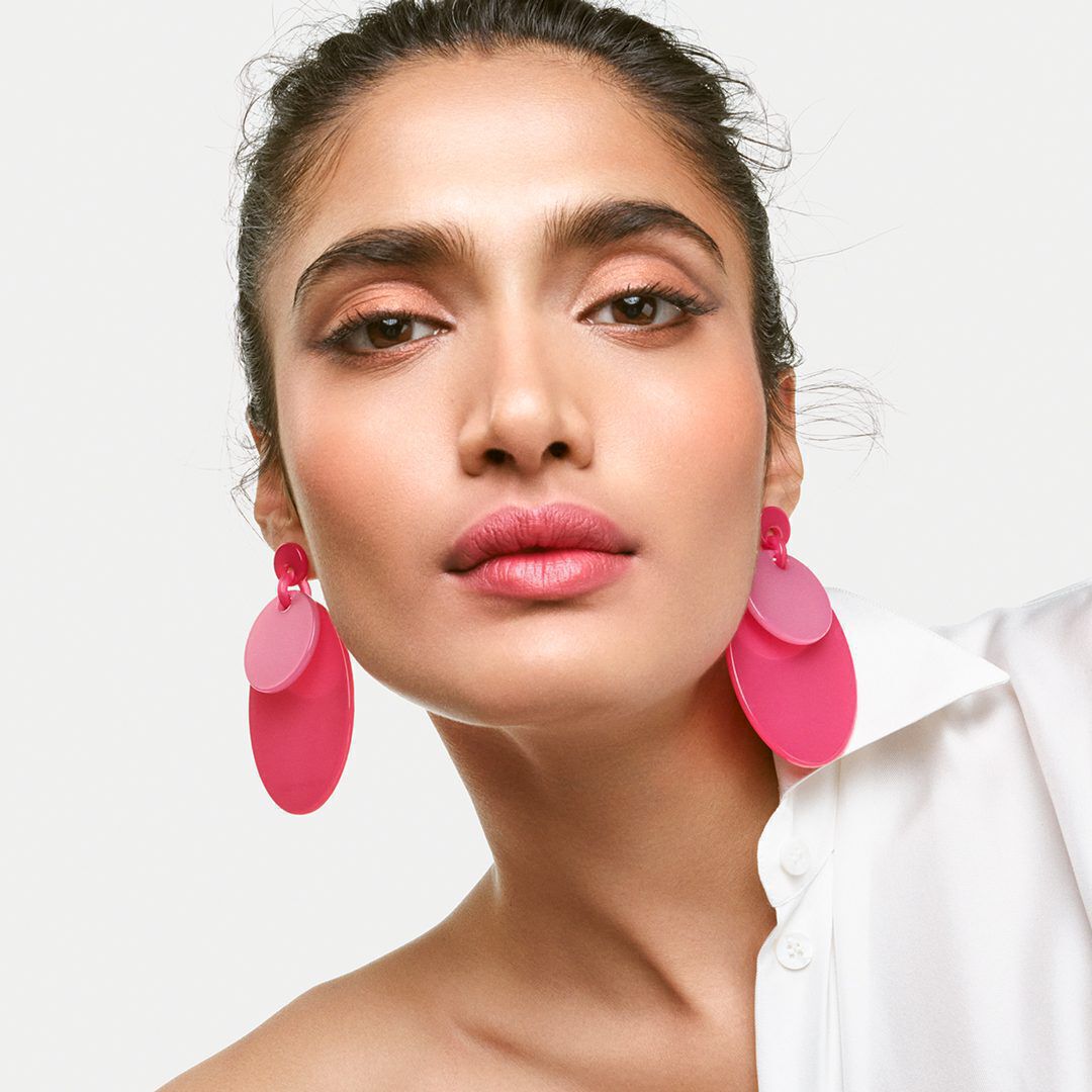 A close-up portrait of a model with a neutral background, featuring striking pink statement earrings and bold lip color, wearing a white shirt. Her expression is confident and engaging.