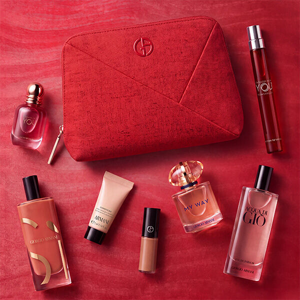 An Armani summer gift set presented on a white marble ledge overlooking a calm blue sea. At the center is a beige knit pouch featuring a blue embroidered GA logo. On the left are Si Passione, Acqua di Gio, a Luminous Silk foundation sample, a liquid blush, and a nude lipstick. On the right are Armani Code, My Way, and Power of You fragrance miniatures.