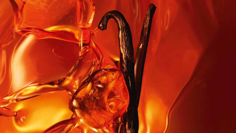Een close-up van twee donkere vanillestokjes en glanzende amberhars tegen een warme, lichtgevende amberkleurige achtergrond.