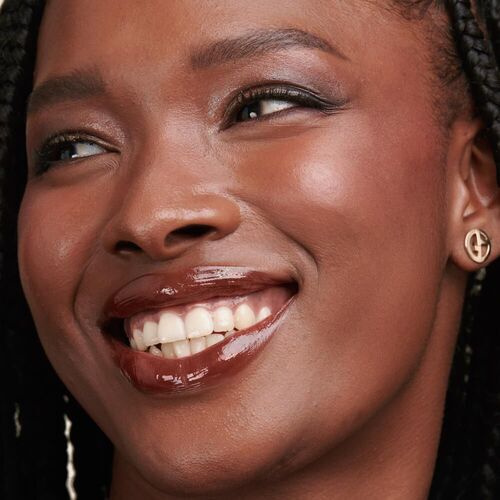 A close-up portrait of a smiling woman with braided hair and subtle makeup, showcasing a glossy lip and earrings.