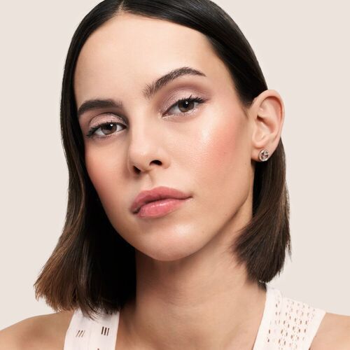 A close-up portrait of a woman with short, straight hair, wearing natural makeup and a white top. She has a neutral expression, and her skin has a smooth, radiant glow against a soft beige background.