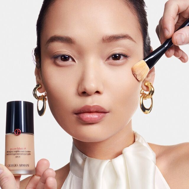 A model showcases a bottle of foundation while a makeup artist applies the product to her cheek with a brush. The background is neutral, and the model has a natural look.