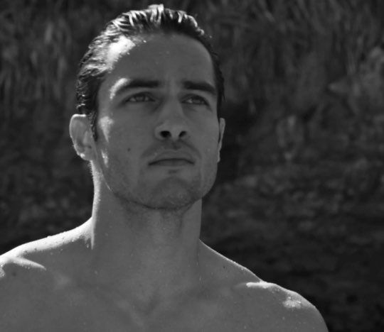 A shirtless man stands with a serious expression, water droplets glistening on his skin, set against a blurred natural background in black and white.