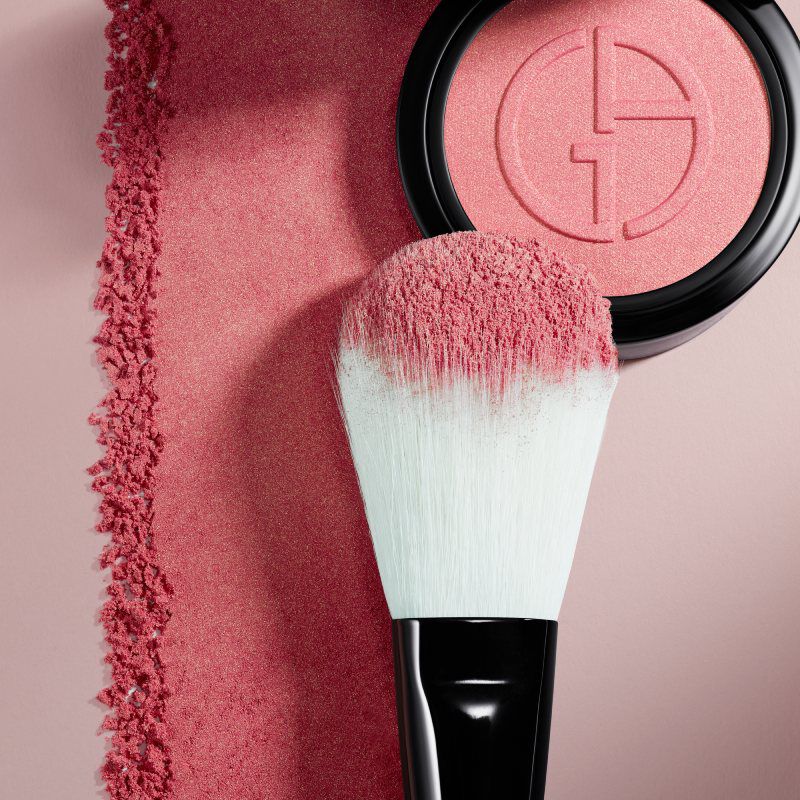 A flat lay image featuring a round blush compact with a pink shade, alongside a makeup brush with a mint green handle and pink bristles, set against a soft pink background. Shimmering blush powder is scattered beside the compact.