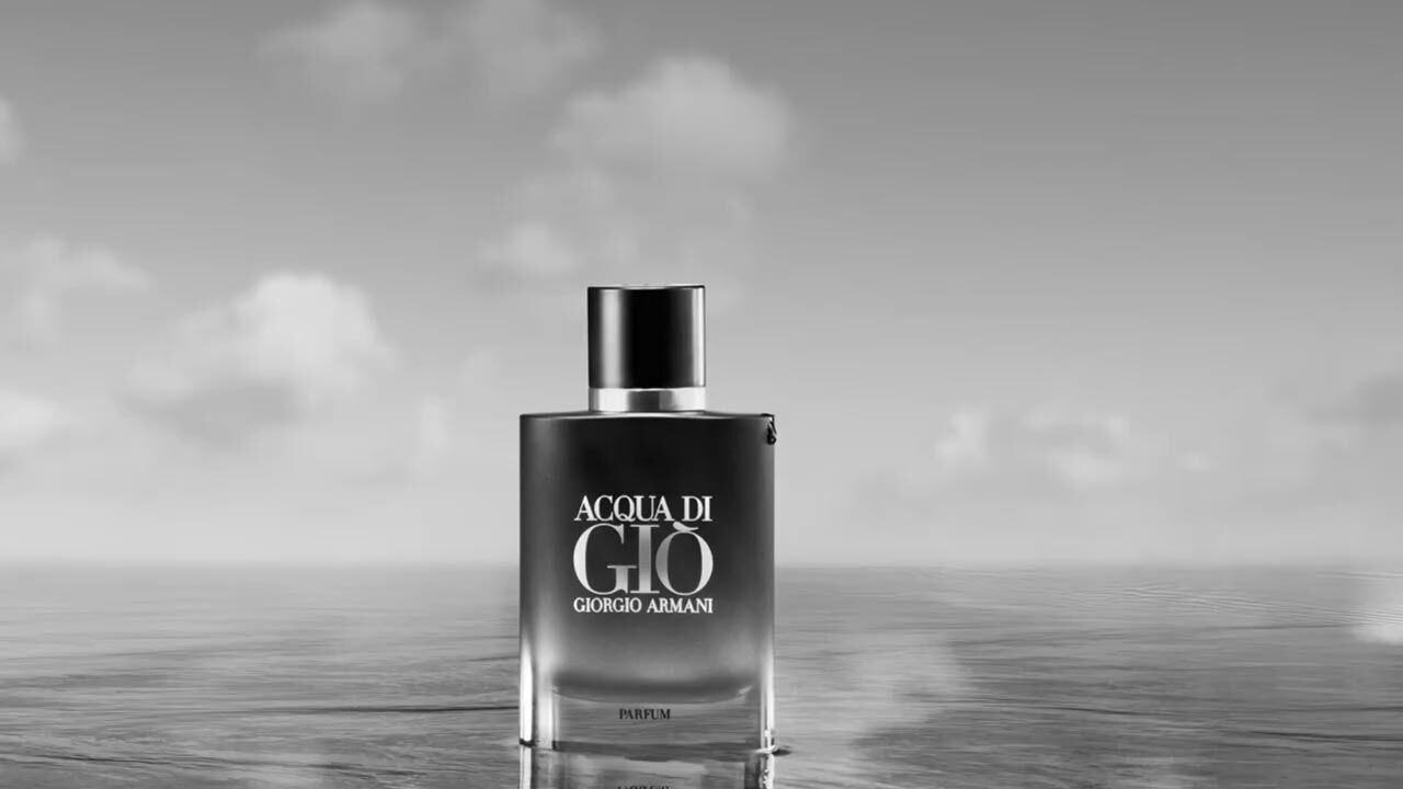 A bottle of Acqua di Gio perfume by Giorgio Armani stands against a serene, cloudy sky and water background. The image is in black and white, emphasizing the elegant design of the bottle.