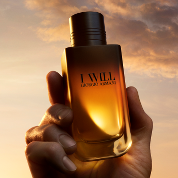 Een hand houdt de I WILL Giorgio Armani-parfumflacon vast tegen een gouden zonsonderganghemel.