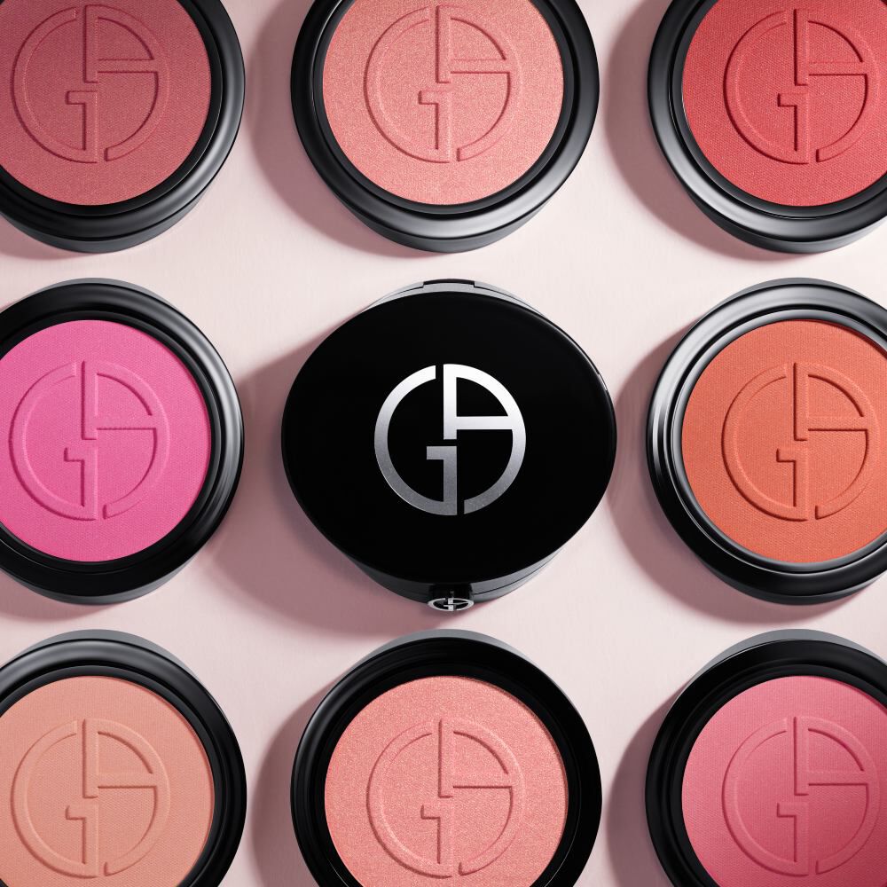 A flat lay image featuring nine blush compacts arranged in a circular pattern, with a central compact displaying a logo. The shades vary from pink to coral, showcasing a range of colors.