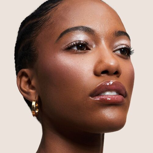 A close-up portrait of a woman with braided hair, showcasing a natural makeup look and glossy lips, set against a neutral background. She wears gold hoop earrings and has a confident expression.