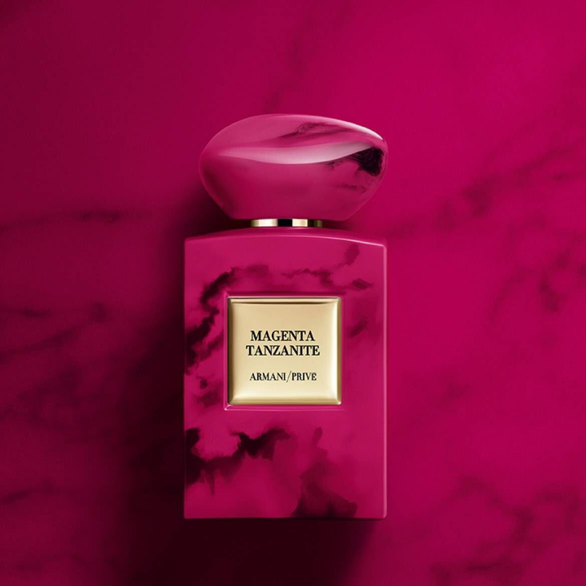 A vibrant pink perfume bottle with a smooth, rounded cap, set against a matching pink background. The label reads "MAGENTA TANZANITE ARMANI/PRIVE."