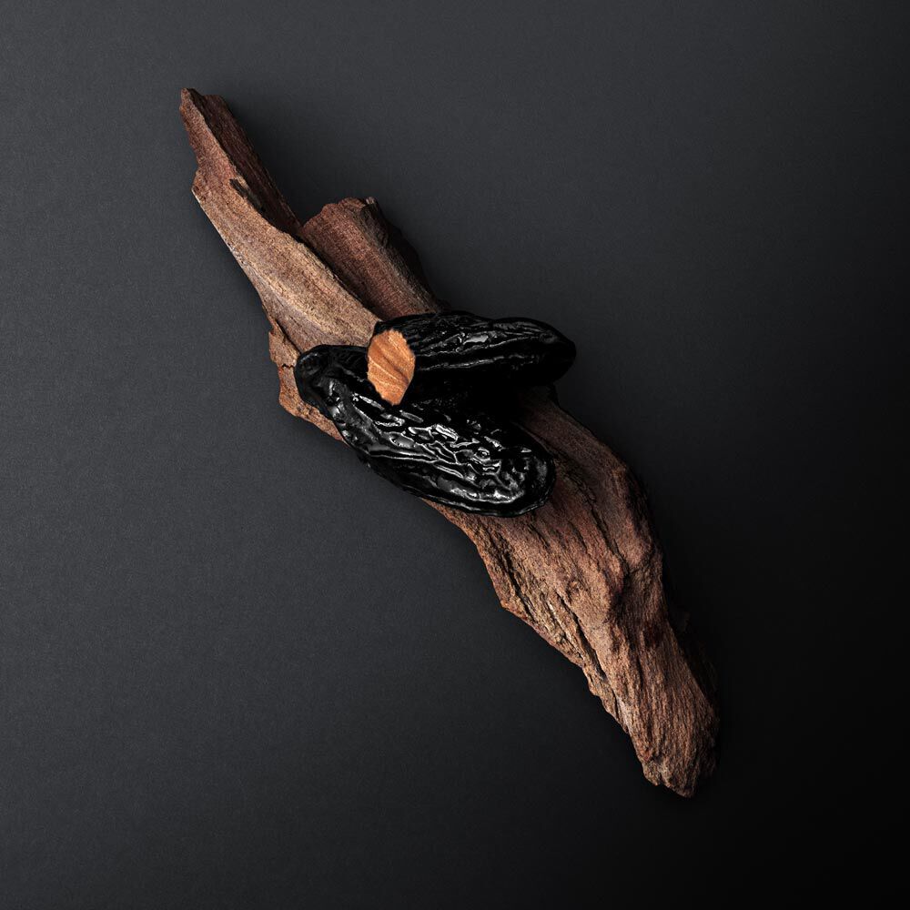 A piece of dark textured wood with two shiny black dried peppers resting on top, set against a black background.