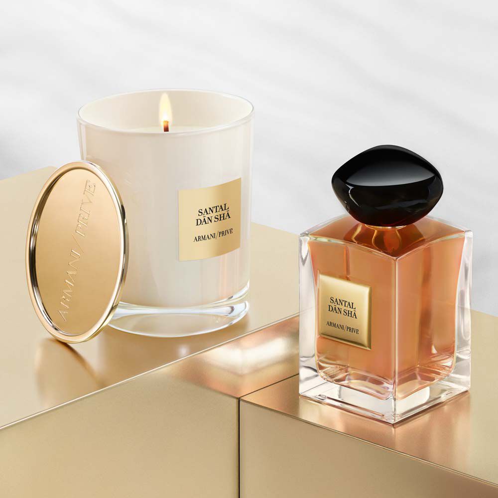 A white candle in a glass holder and a rectangular perfume bottle, both featuring gold accents and labeled "Santal Dan Sha" by Armani Prive, are displayed on a sleek, metallic surface.