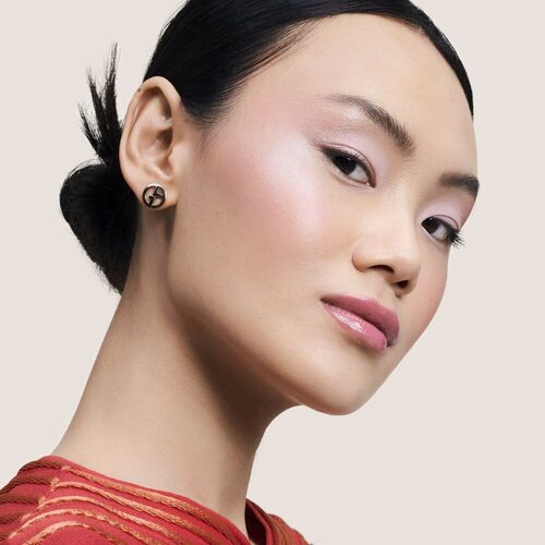 A woman with a sleek bun and wearing subtle makeup poses confidently, showcasing a pair of stylish earrings. She is dressed in a red garment with a textured pattern, against a neutral background.