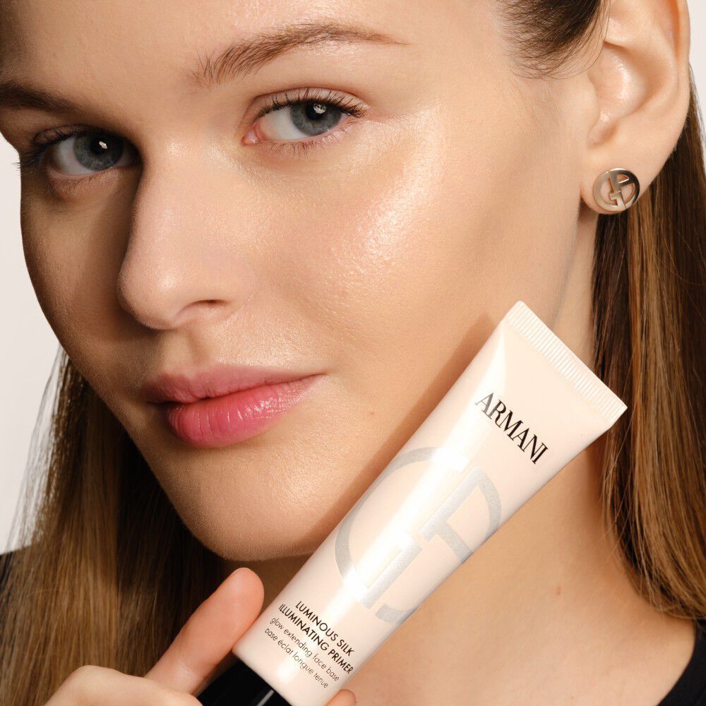 A face close-up with a hand that holds a beige tube of makeup primer near the cheek; long brown hair; clean, minimal aesthetic.