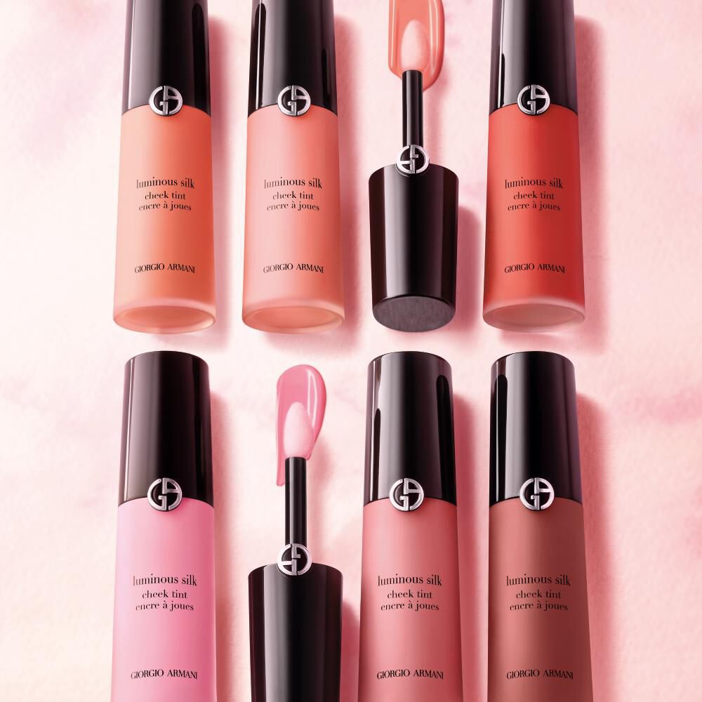 An arrangement of Giorgio Armani Luminous Silk Cheek Tint bottles in various peach, pink, and nude shades, displayed against a soft pink background, with a small applicator in the foreground.