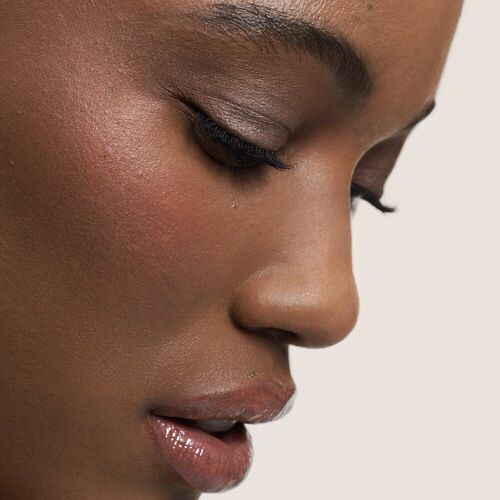 Close-up profile of a person with smooth skin and elegant makeup, featuring prominent cheekbones and soft lips. The background is a light neutral color.