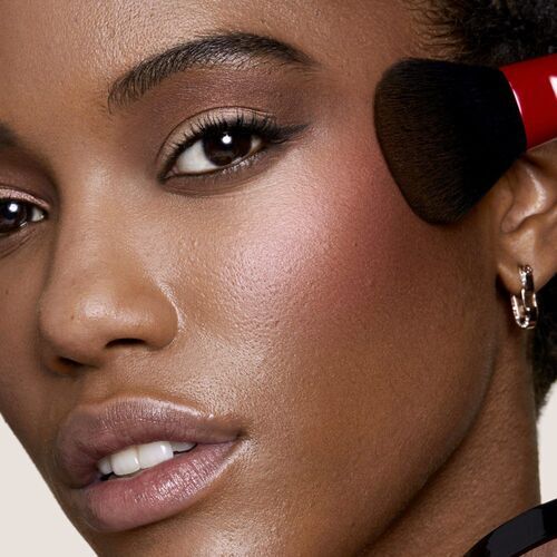 A close-up of a woman applying blush to her cheek with a makeup brush, showcasing her natural beauty and glowing skin.