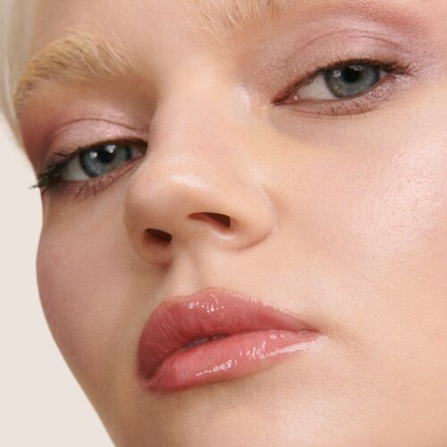 Close-up of a person’s face, showcasing natural makeup with soft pink tones and glossy lips, highlighting their blue eyes and light-colored eyebrows.