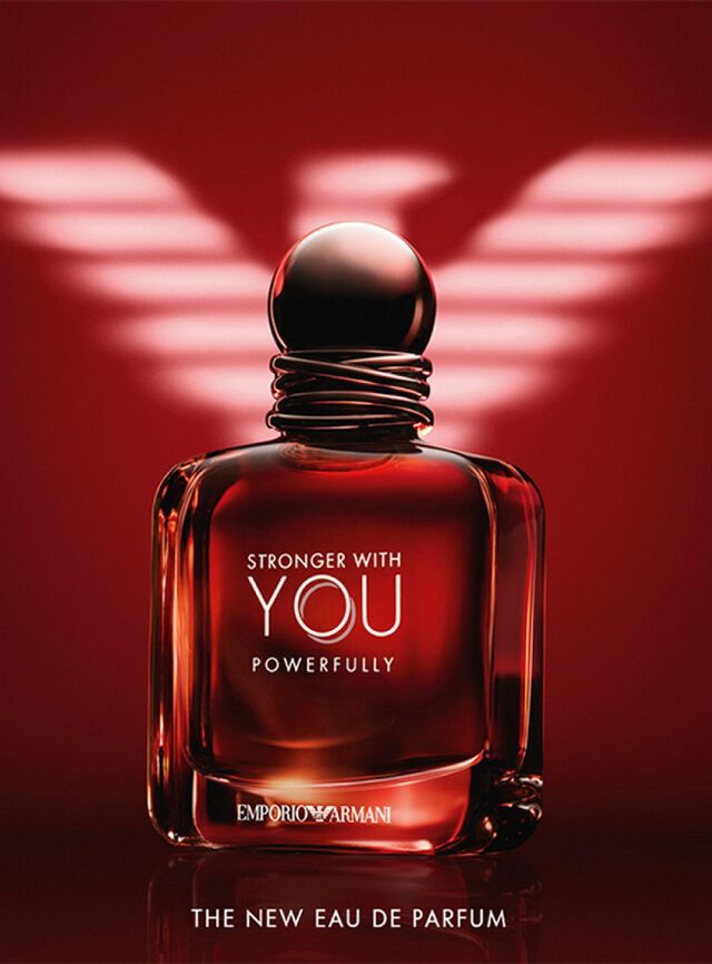 Une publicite spectaculaire pour l'Eau de Parfum « Stronger With You Powerfully » d'Emporio Armani. L'image presente un flacon en verre carre et epais, rempli d'un liquide rouge ambre intense qui semble briller de l'interieur. Le flacon possede un bouchon sombre et arrondi avec un detail de fils entrelaces autour du col. Les inscriptions « STRONGER WITH YOU POWERFULLY » et « EMPORIO ARMANI » sont imprimees en argent et blanc. En arriere-plan, un grand logo de l'aigle Armani stylise est illumine en blanc sur un fond rouge profond. En bas, le texte indique « THE NEW EAU DE PARFUM ».