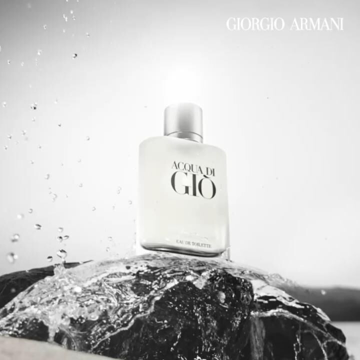 A bottle of Acqua di Gio by Giorgio Armani sits on a rock, surrounded by splashing water, with a minimalist black and white aesthetic.