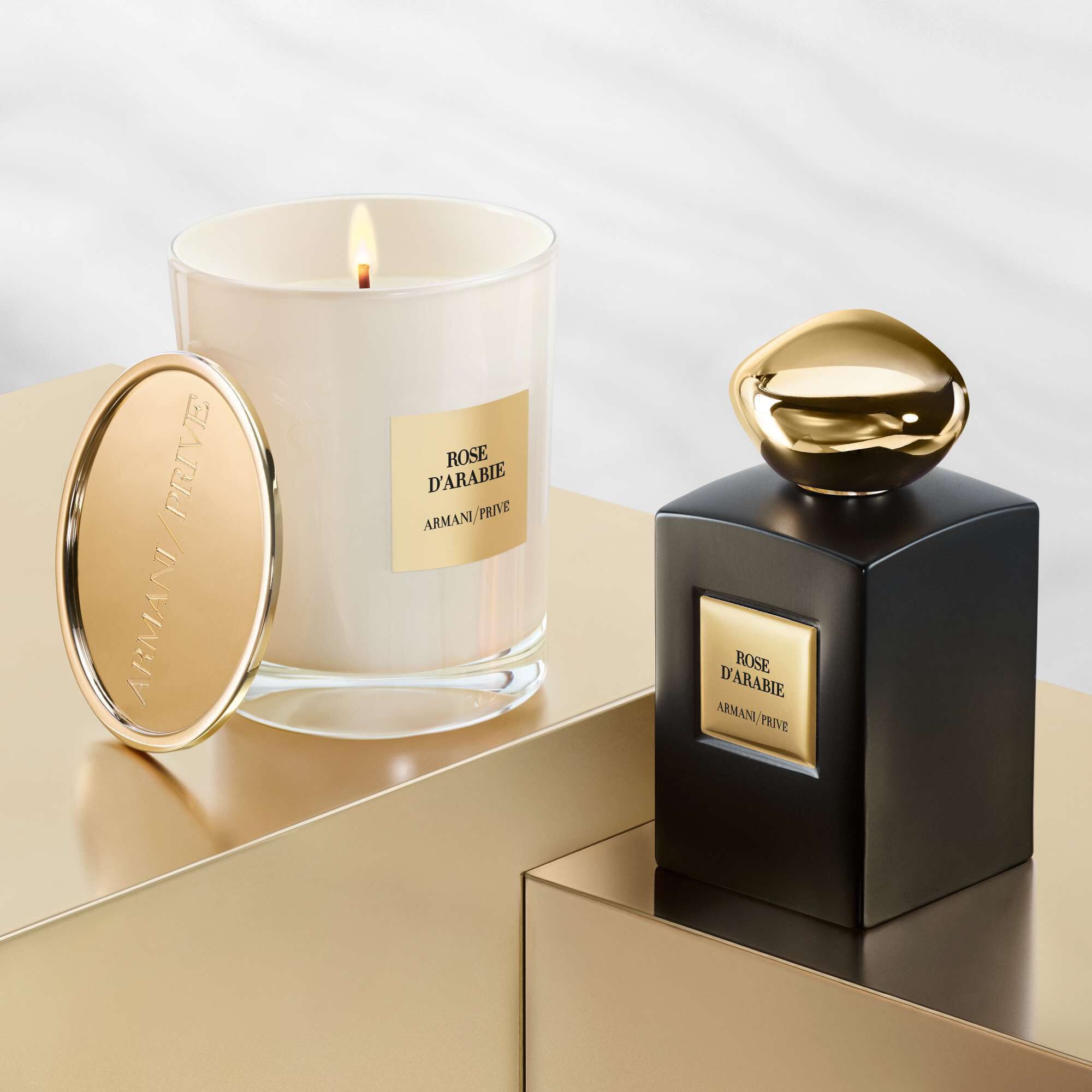 Alt text: A luxurious candle and perfume bottle both labeled "Rose d'Arabie" by Armani Prive, displayed on golden surfaces with a soft, blurred background. The candle is in a glass holder topped with a gold lid, while the perfume bottle is sleek and black with a gold cap.