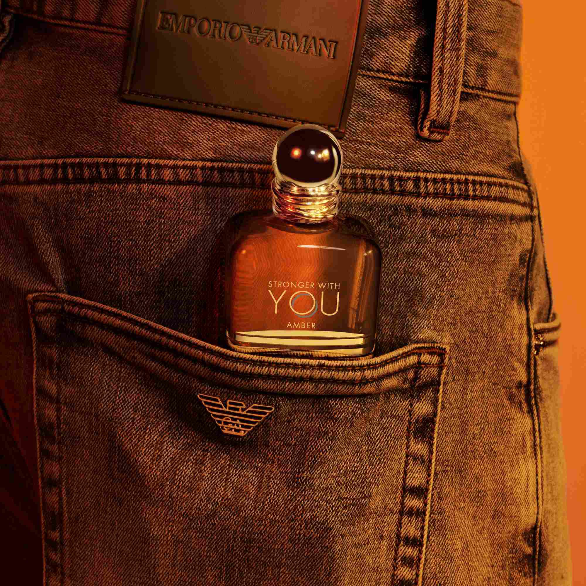 Een Emporio Armani Stronger With You Amber parfumfles die uit de achterzak van een spijkerbroek steekt, met een donkere leren