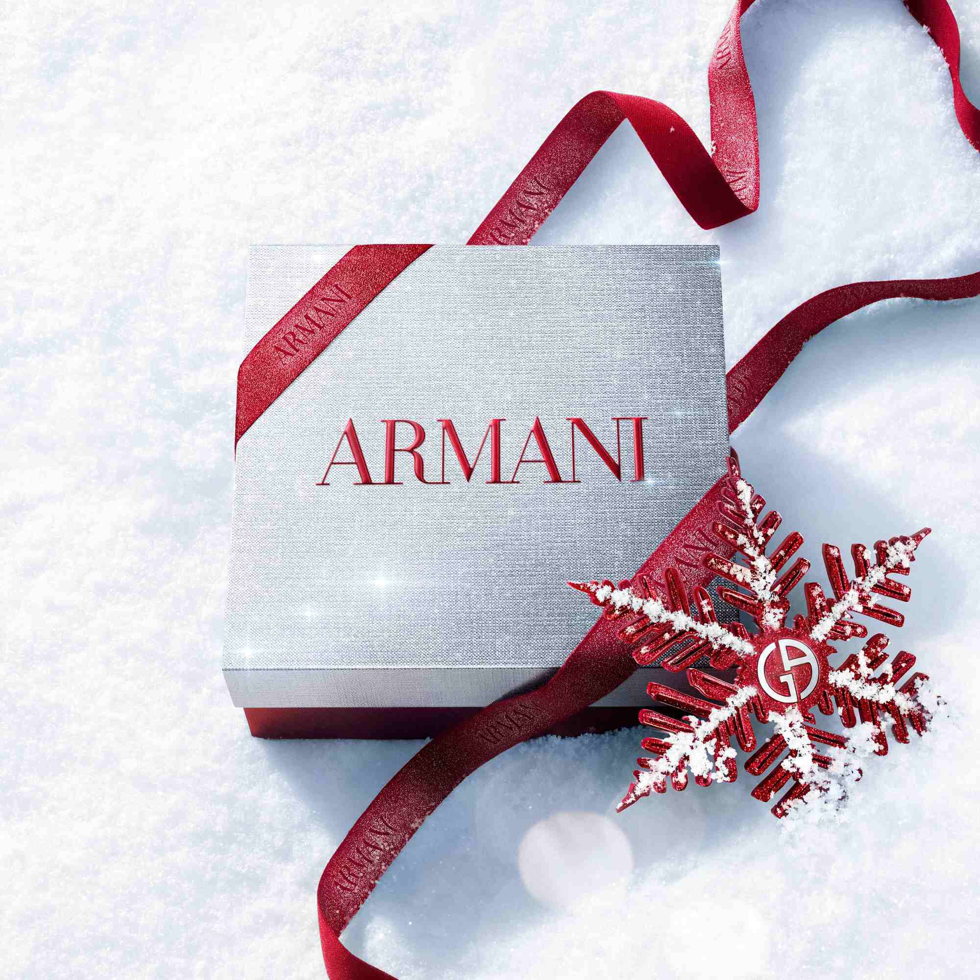 Zilveren Armani Glamourous Winter Holiday Cadeaudoos met rood glitterlint en Giorgio Armani rode sneeuwvlok versiering op een fonkelend besneeuwd oppervlak — feestelijke luxe geurverpakking.