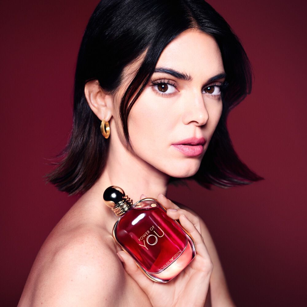 Kendall Jenner houdt de ARMANI POWER OF YOU-parfumfles dicht tegen haar borst, tegen een diepe robijnrode achtergrond.