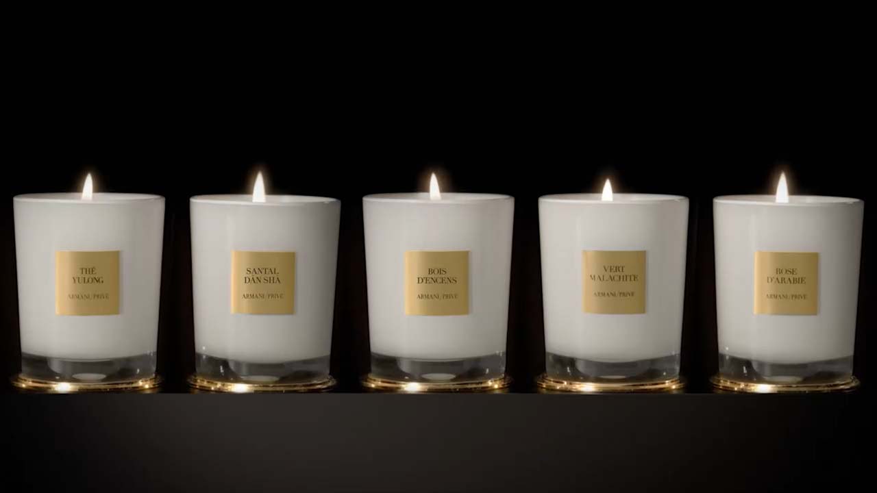 Cinq bougies blanches allumées sur fond noir, étiquetées avec des noms de parfums élégants.
